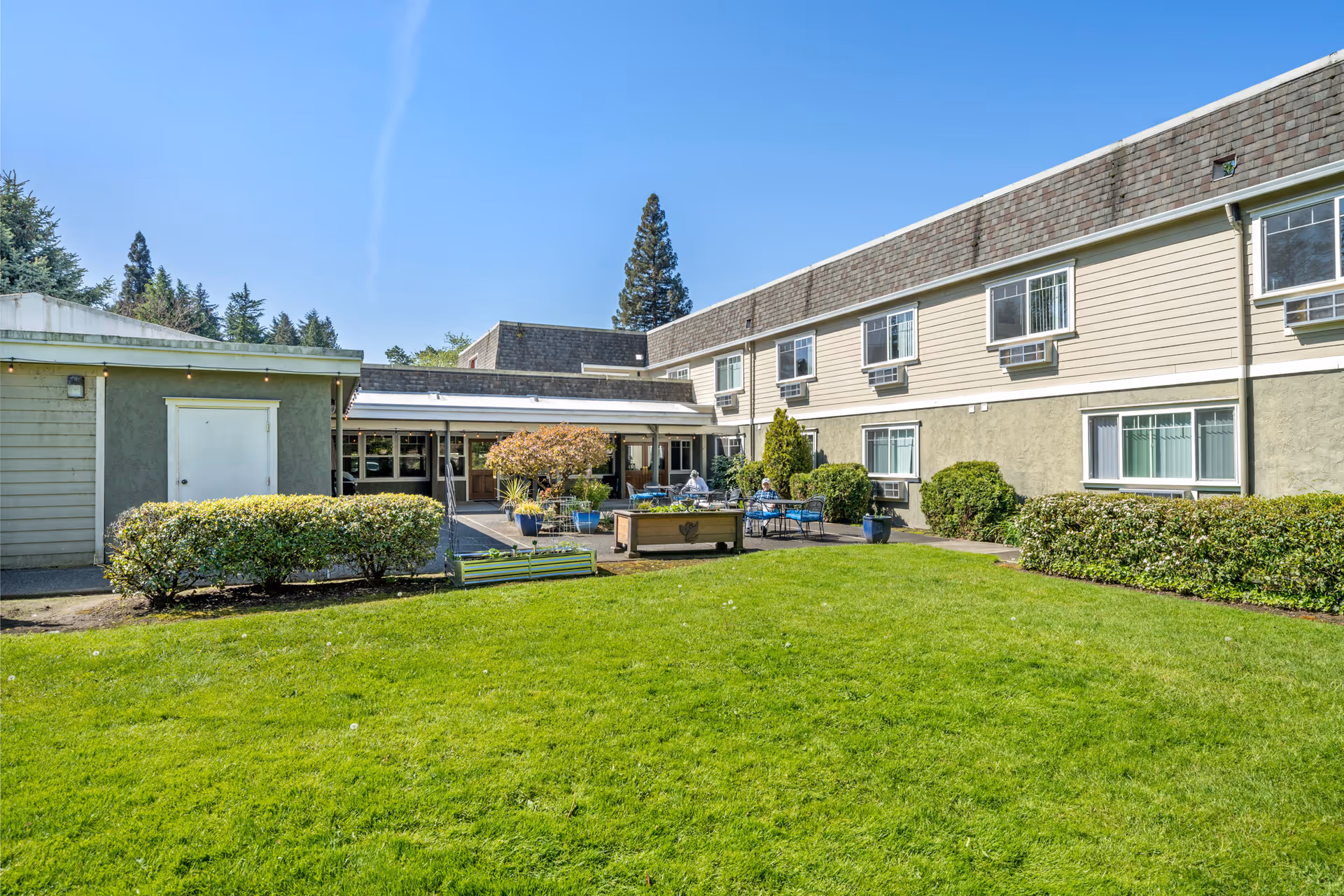 Green lawn and patio seating in front of a two-story beige senior living building under a clear blue sky.