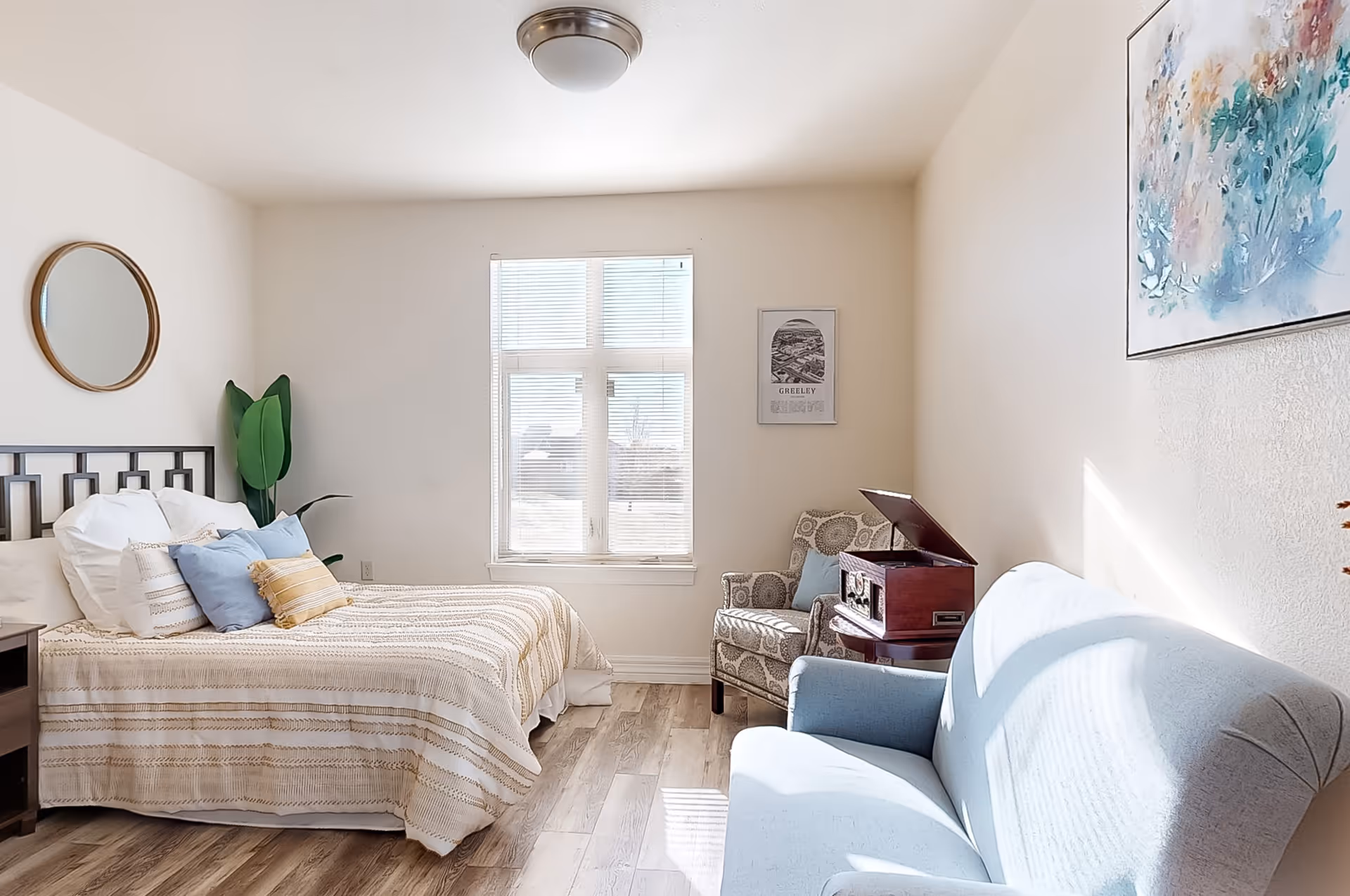Sunlit bedroom with a made bed, bedside table, patterned armchair with a record player, and a blue loveseat.