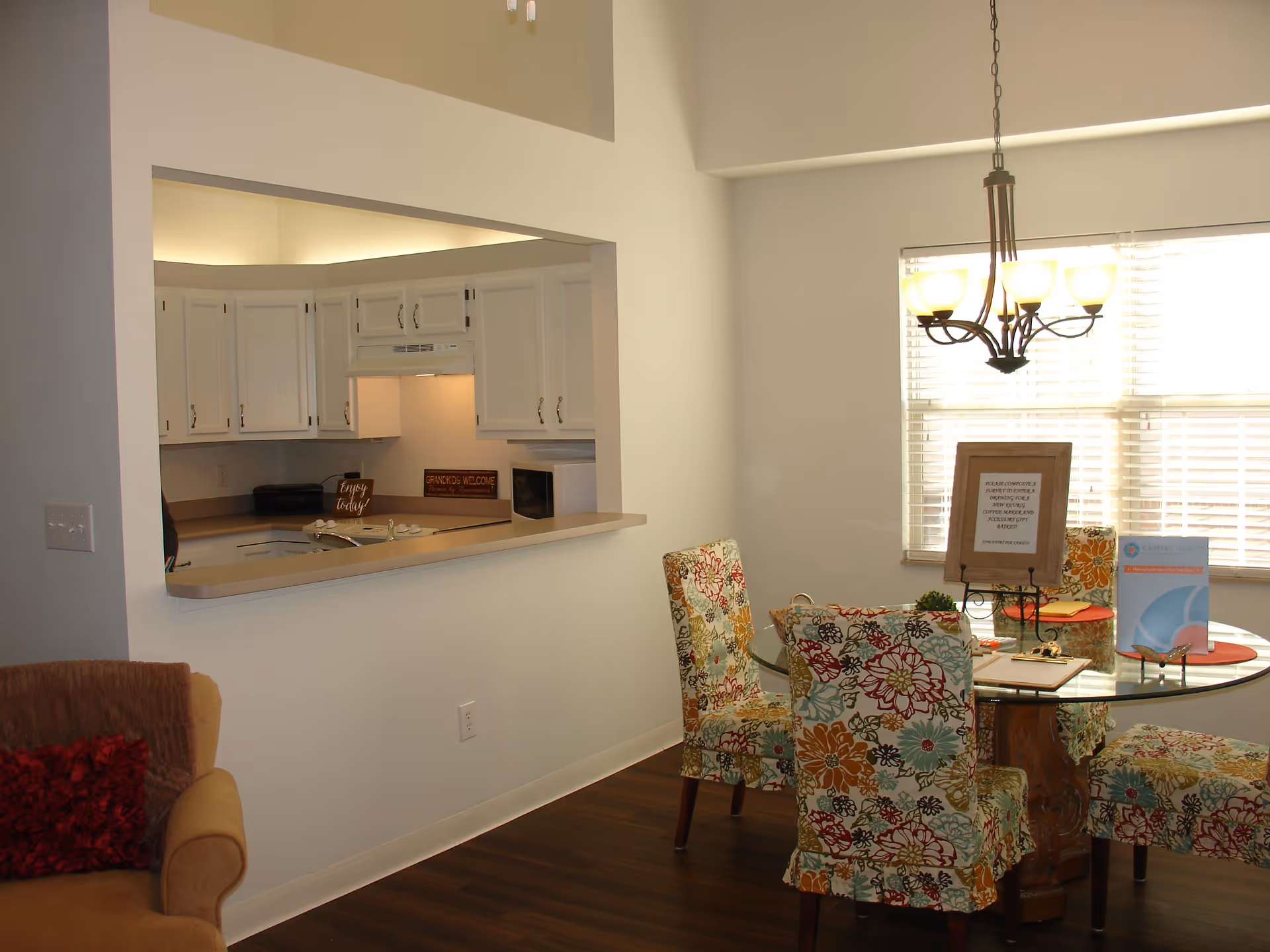 Interior view of a dining area and kitchen in a senior living facility. The dining area features a round glass table with four floral-patterned chairs. A chandelier with five lights hangs above the table. The kitchen is visible through a pass-through window and has white cabinets, a microwave, and a countertop with a sink. There is a beige armchair with a red pillow partially visible on the left side.