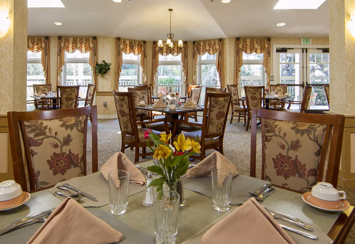 A well-lit dining room with multiple tables set for a meal. Each table has floral-patterned chairs, folded beige napkins, glassware, and a small vase with colorful flowers. Large windows with decorative curtains allow natural light to fill the room, and a chandelier hangs from the ceiling.