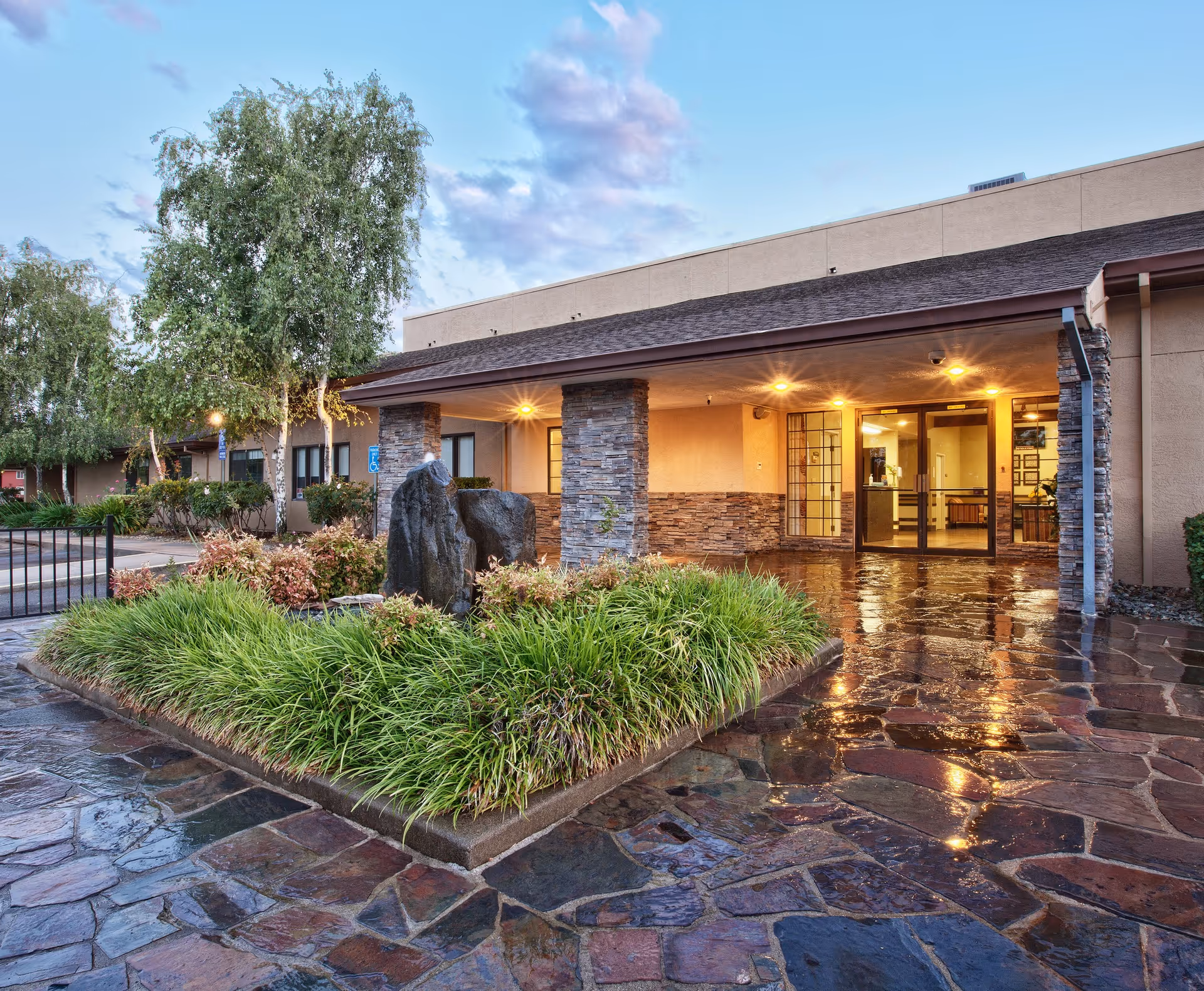 Entrance of Oak River Rehab facility with a covered porch supported by stone pillars, a stone-paved walkway, and landscaped greenery including bushes and a tree. The building exterior is beige with large glass doors and windows, illuminated by warm lights.