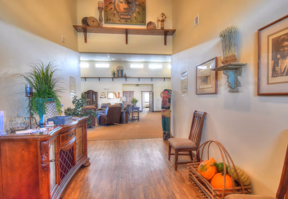 Interior view of a senior living facility hallway leading into a spacious living room area. The hallway has wooden flooring, a wooden sideboard with plants and decorative items on the left, and a chair with a basket of pumpkins and gourds on the right. The living room area has comfortable seating, plants, and wall decorations with high windows letting in natural light.