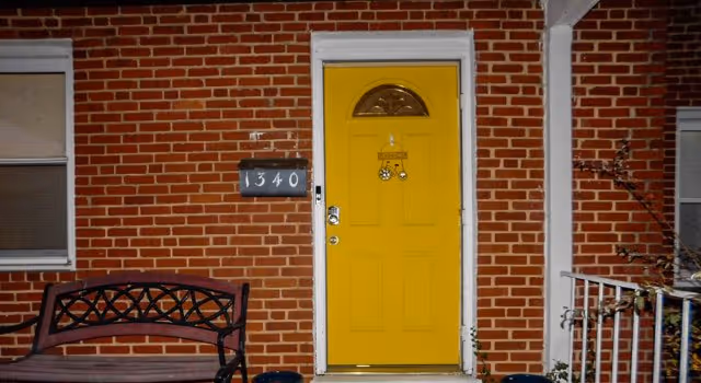 Front entrance of a brick building with a bright yellow door, a black bench to the left, a house number plaque displaying 1340, and a white railing on the right side.