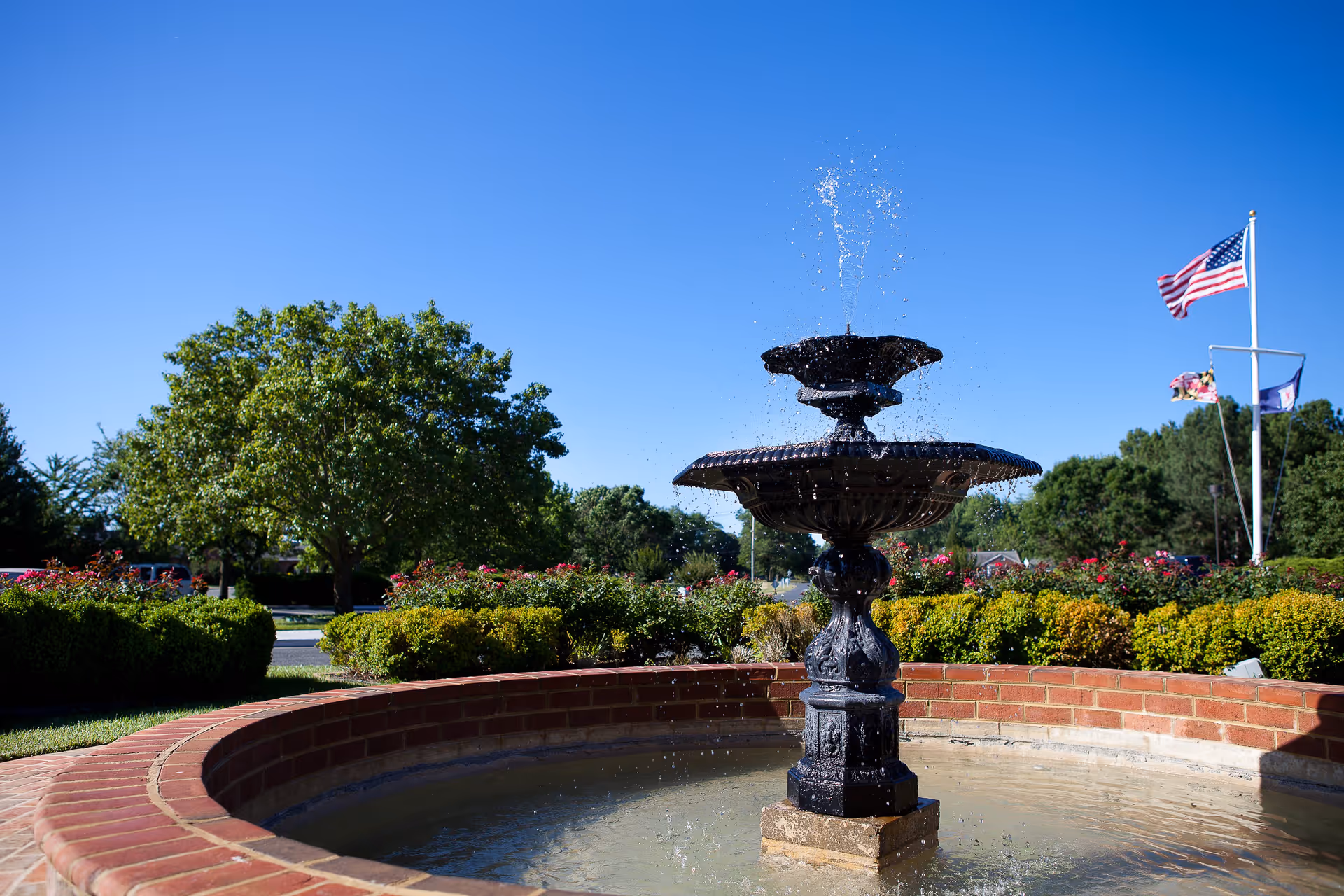A black decorative water fountain in a circular brick basin with water spraying upwards, surrounded by green bushes and trees under a clear blue sky. Three flags, including the American flag, are visible in the background.