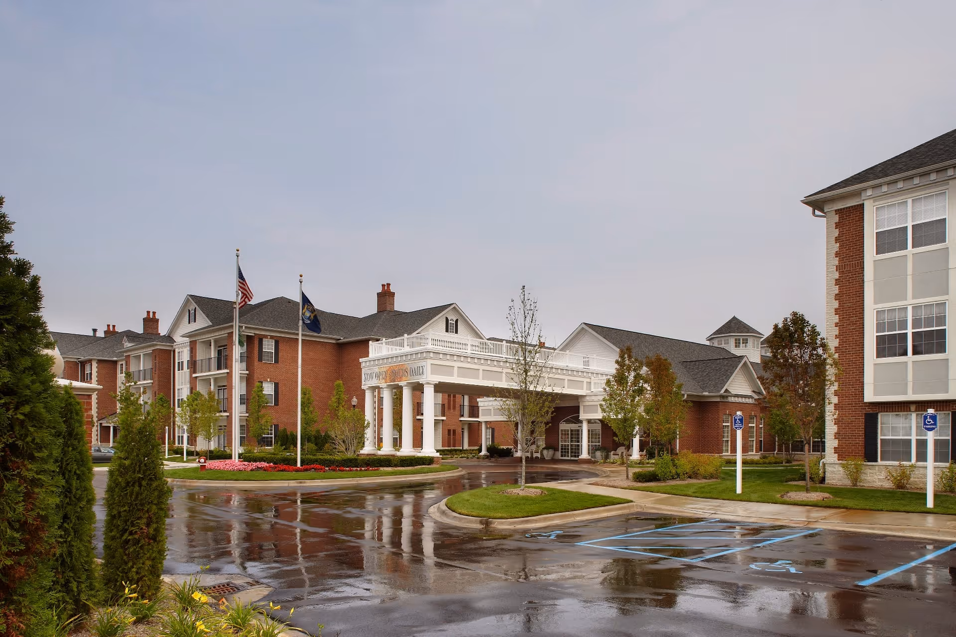 Exterior view of a senior living facility with red brick buildings, white columns at the entrance, two flagpoles with flags, landscaped greenery, and a wet parking lot with handicap parking spaces.