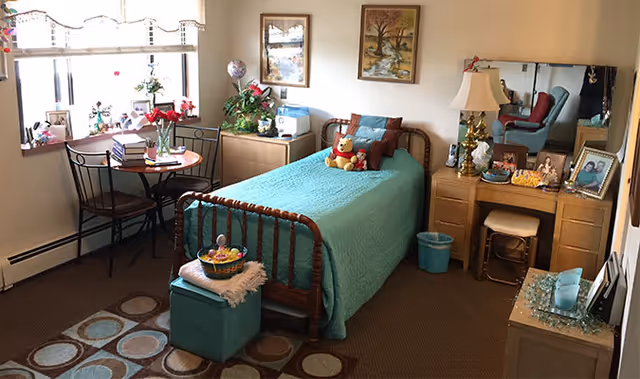 A cozy bedroom in an assisted living facility with a single bed covered in a teal quilt and a teddy bear on the pillow. There is a small round table with two chairs near a window with blinds, a wooden dresser with a lamp and mirror, and various decorative items including framed pictures, plants, and a basket on a teal ottoman at the foot of the bed.