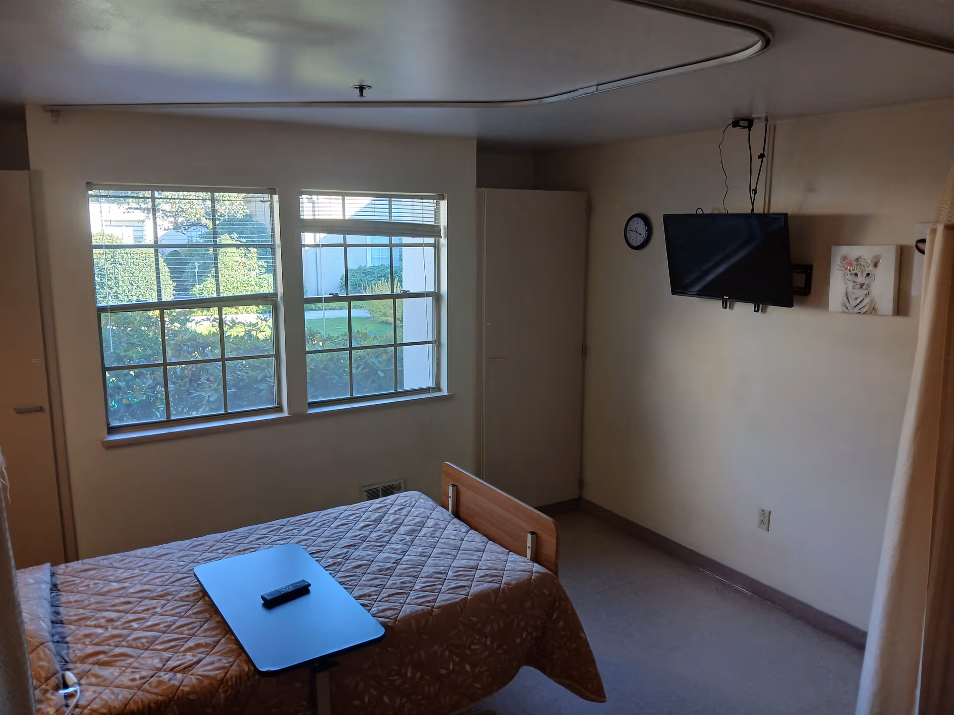 A small resident bedroom with a quilted bed, overbed table and remote, large windows showing greenery, a wall-mounted TV and a closet.