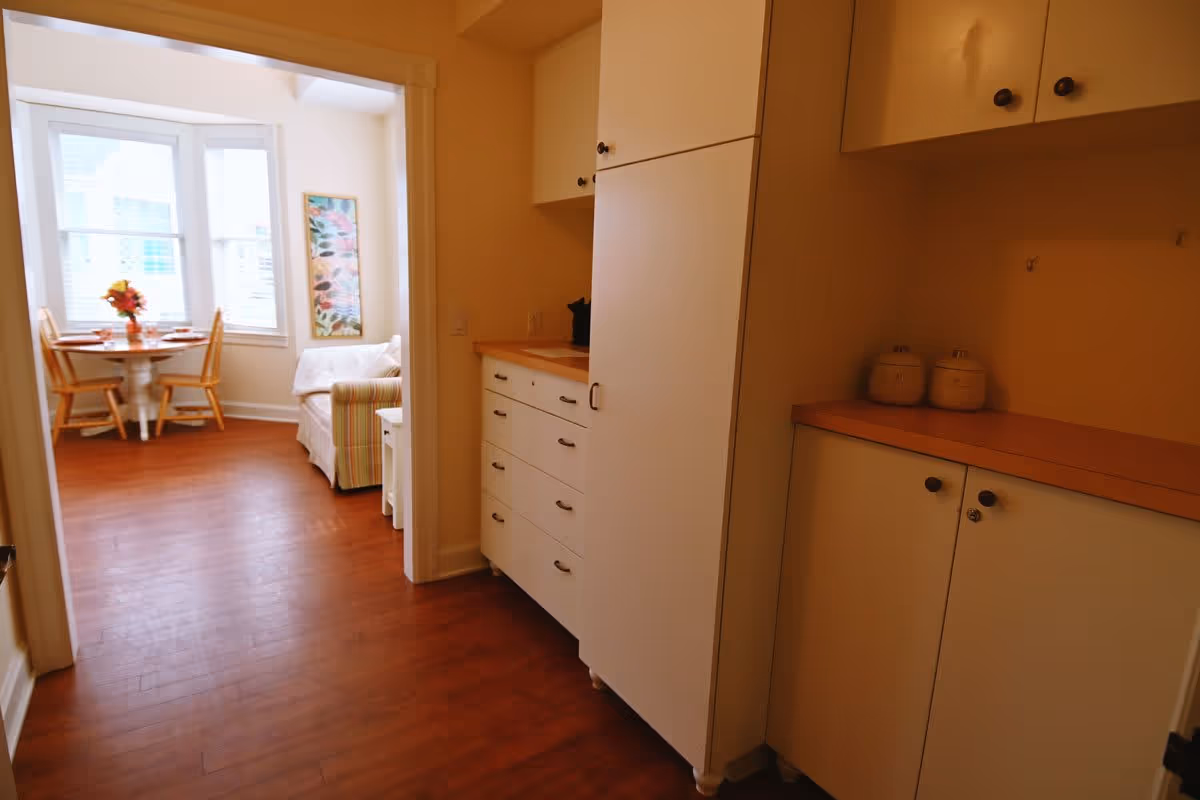 View of a hallway with white cabinets and wooden countertops leading into a bright room with a round dining table, two wooden chairs, a small couch, and a large window with blinds. A colorful vertical painting is on the wall near the window.