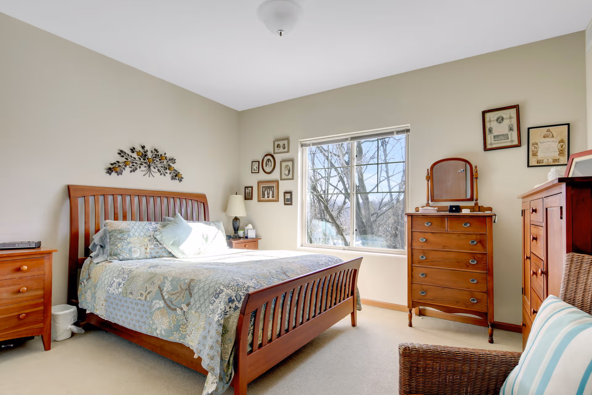 A bright bedroom with a wooden bed frame and a patterned quilt. There are two wooden nightstands with a lamp and some items on them. A window lets in natural light and shows a view of trees outside. The walls have framed pictures and decorative items. There is also a wooden dresser with a mirror and another wooden cabinet in the room.