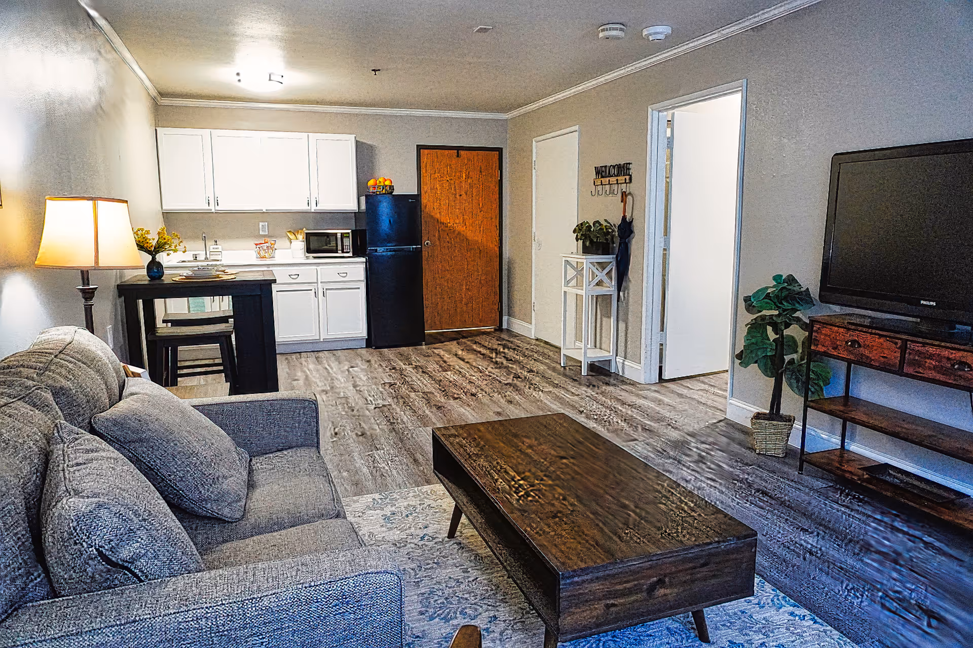 A cozy living room and kitchenette area featuring a gray sofa with cushions, a wooden coffee table on a patterned rug, a TV on a wooden stand, a small dining table with two chairs, white kitchen cabinets, a black refrigerator, and a wooden door. The room has light-colored walls and wood flooring, with a small plant and a welcome sign near the doorway.