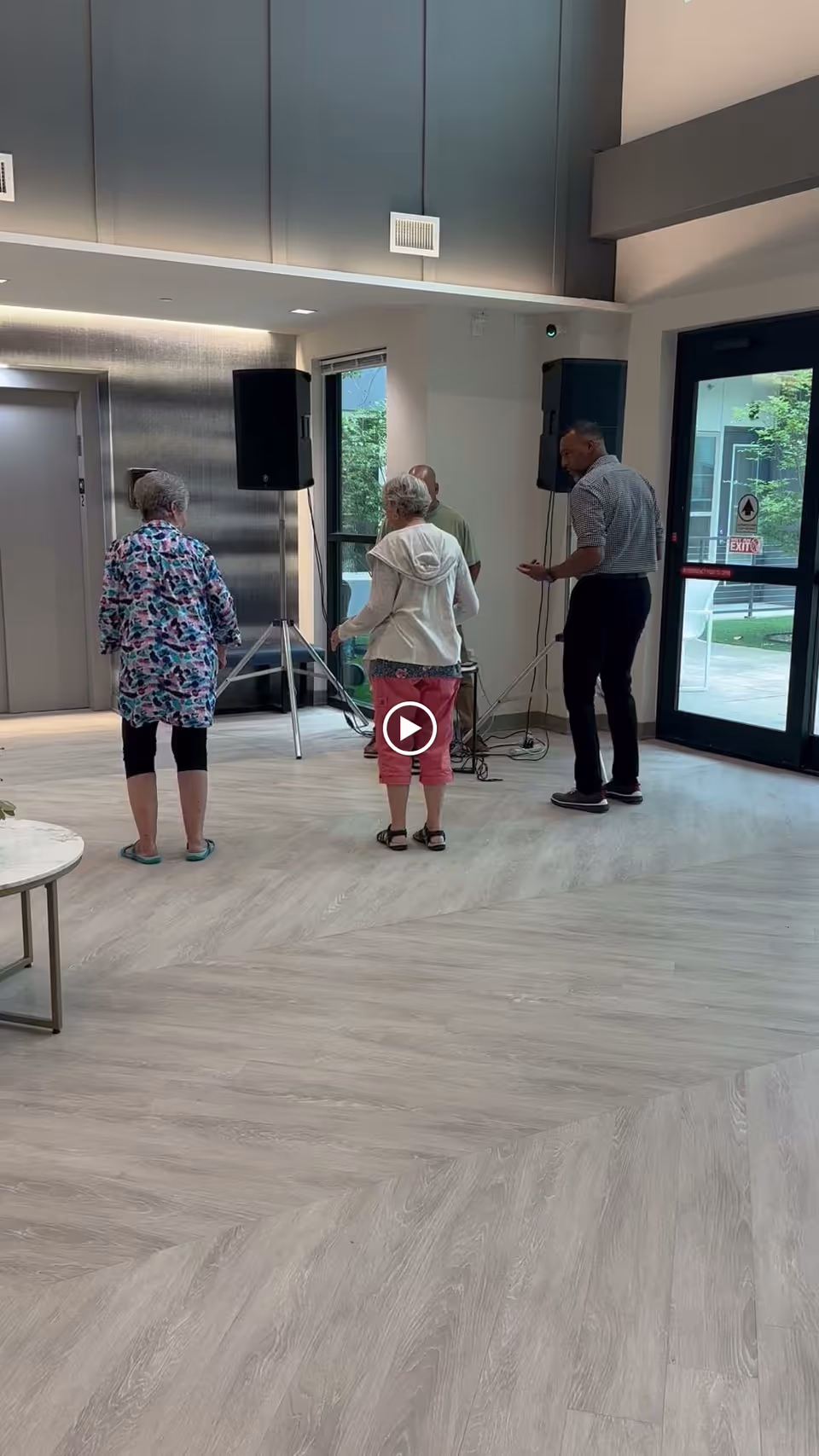 Four people standing in a modern indoor common area with light wood flooring, large windows, and a glass door leading outside. Two large speakers on stands are positioned near the windows, and there is an elevator door on the left side of the image.