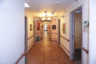 A long, well-lit hallway in a senior living facility with tiled floors and handrails on both sides. The walls are decorated with framed artwork, and there are several doors along the corridor. Ceiling lights and a chandelier provide illumination.