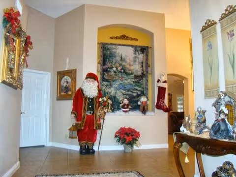 Interior view of a living area decorated for Christmas with a large Santa Claus figure, a poinsettia plant, Christmas stockings, and various holiday figurines on a wooden table. The walls are adorned with framed artwork and a large tapestry depicting a garden scene.