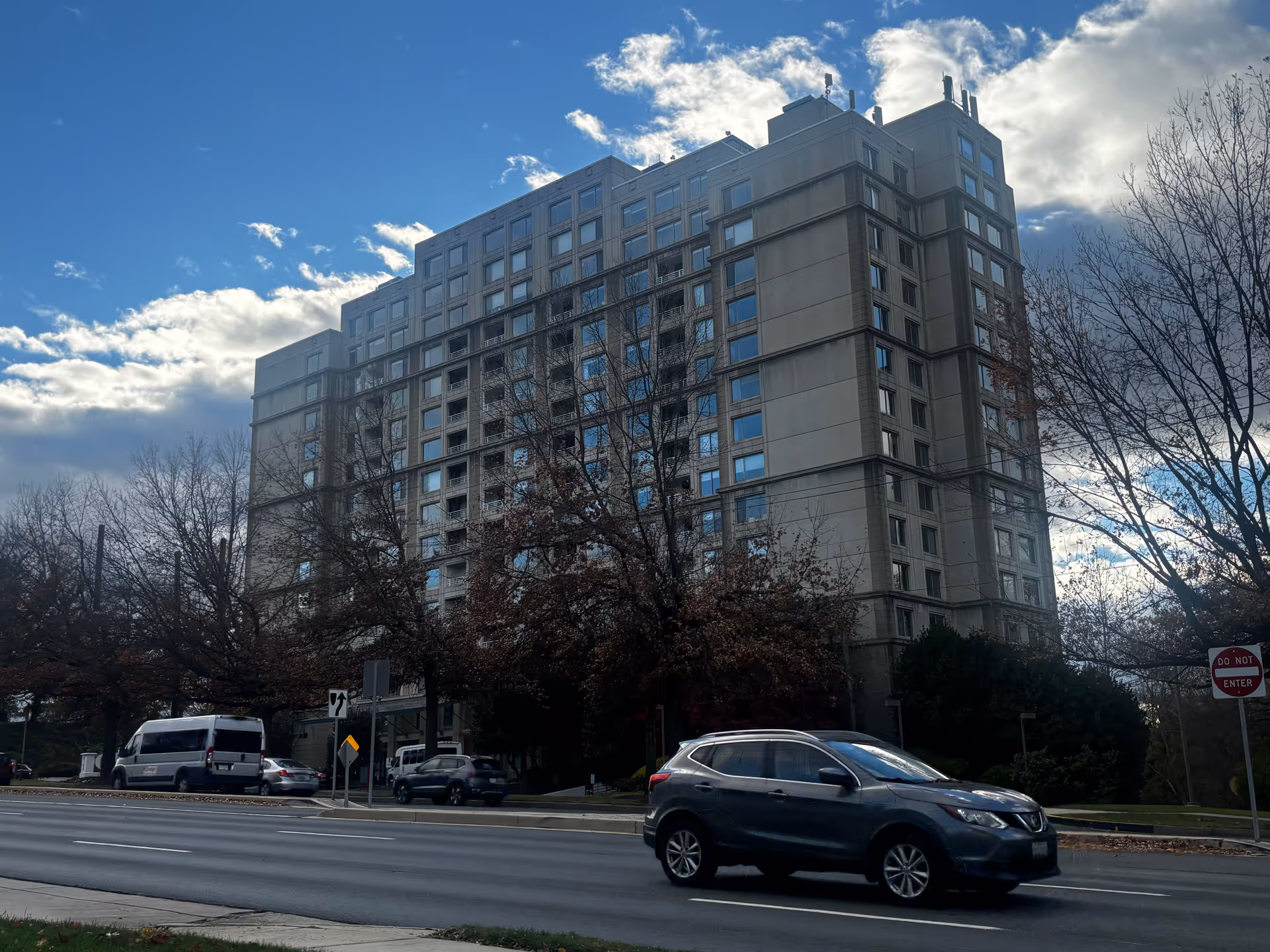 Exterior view of a multi-story residential building with many windows, surrounded by leafless trees and a few cars driving on the road in front. The sky is partly cloudy with patches of blue.