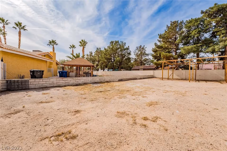 Outdoor area with a sandy ground, a small gazebo, some palm trees, and a yellow building on the left side. There is a wooden frame structure on the right side and a white block wall enclosing the area.