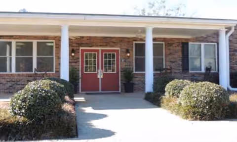 Brick building entrance with red double doors, white columns, and trimmed shrubs.