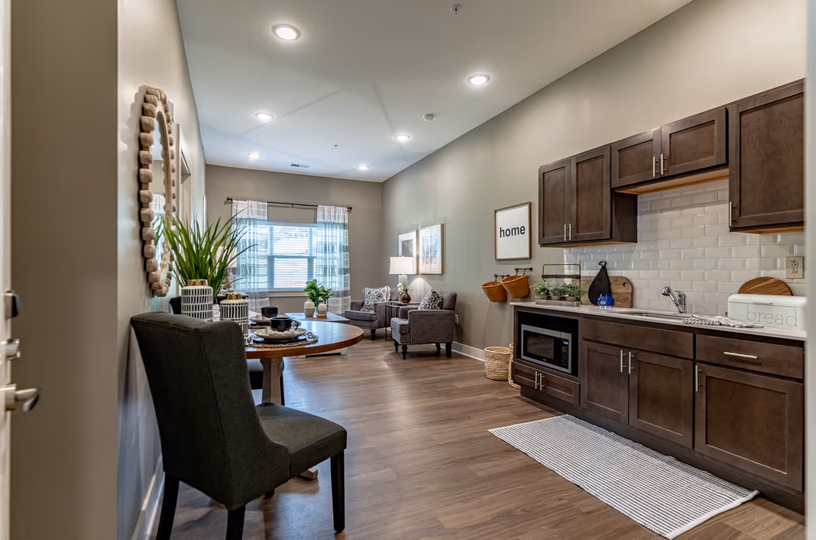 A modern, open-concept living space in a senior living facility featuring a small kitchen area with dark wood cabinets, a microwave, and a sink on the right. A striped rug lies in front of the kitchen. On the left, there is a round dining table with a dark upholstered chair and decorative plants. In the background, a cozy sitting area with two armchairs, a side table with a lamp, and framed artwork on the wall is visible. A window with sheer curtains allows natural light into the room.