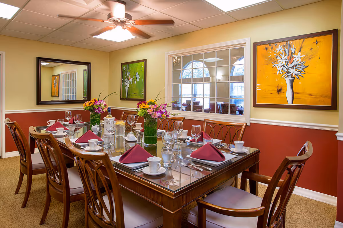 A dining room with a rectangular wooden table set for eight people. The table has glass top and is arranged with white cups, saucers, glasses, silverware, and burgundy folded napkins. Two vases with colorful flower arrangements are placed in the center. The room has beige and red walls with two framed floral paintings and a large mirror. A ceiling fan with lights is mounted on the white ceiling. A window with multiple panes looks into another room with arched windows.