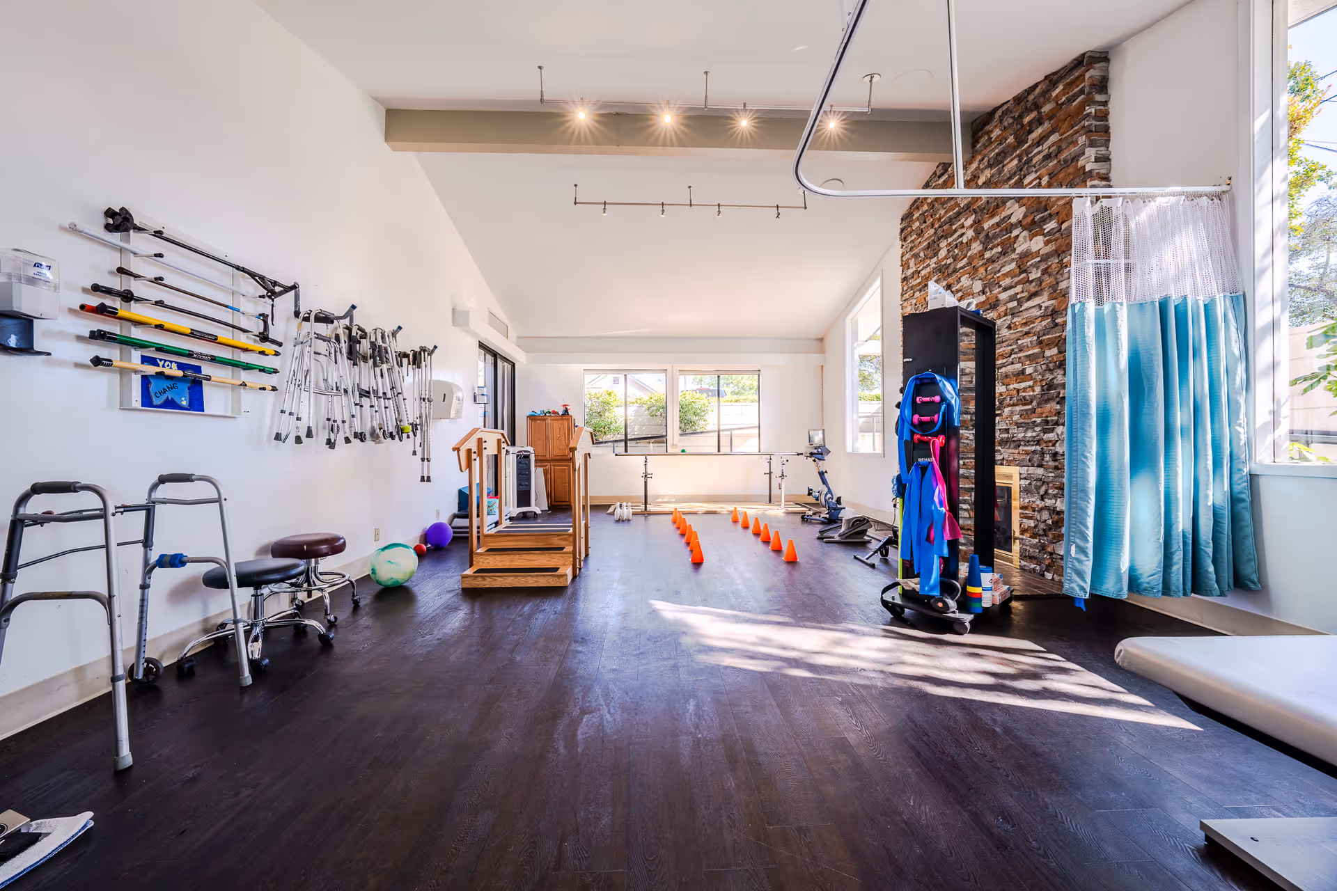 A spacious rehabilitation or physical therapy room with various mobility aids such as walkers, crutches, and canes hanging on the wall and standing on the floor. The room features exercise equipment including a stationary bike, therapy balls, and orange cones arranged in a line for agility training. There is a wooden step platform and a curtain divider near large windows letting in natural light. The room has a dark wooden floor and a stone accent wall.