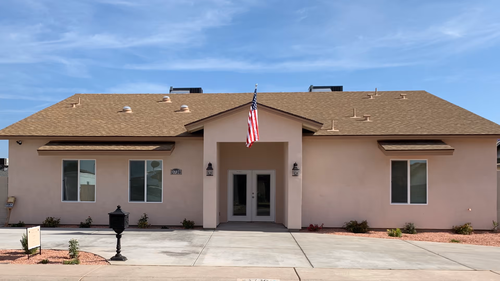Front exterior of a single-story assisted living building with an American flag over the entrance and a paved driveway.