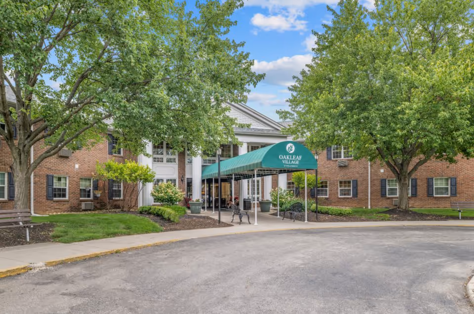 Front entrance of Oakleaf Village of Columbus showing a brick building with white columns, a green canopy, benches and surrounding trees.