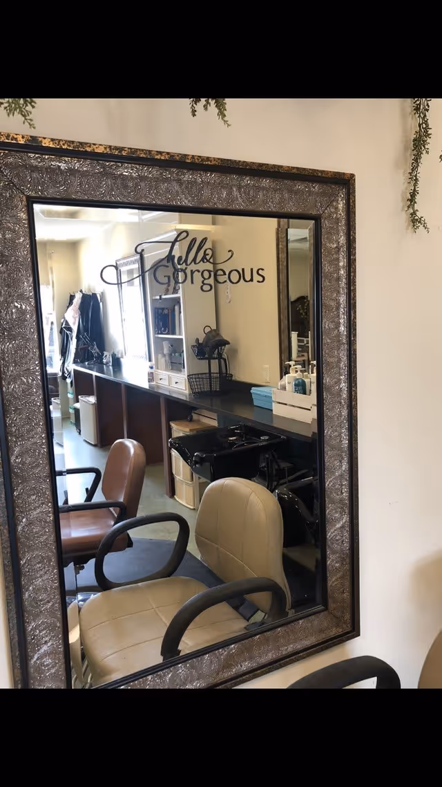 A large ornate framed mirror on a wall reflecting a salon area with chairs, a counter with various items including hand sanitizer, and a sign on the mirror that says 'hello Gorgeous'.