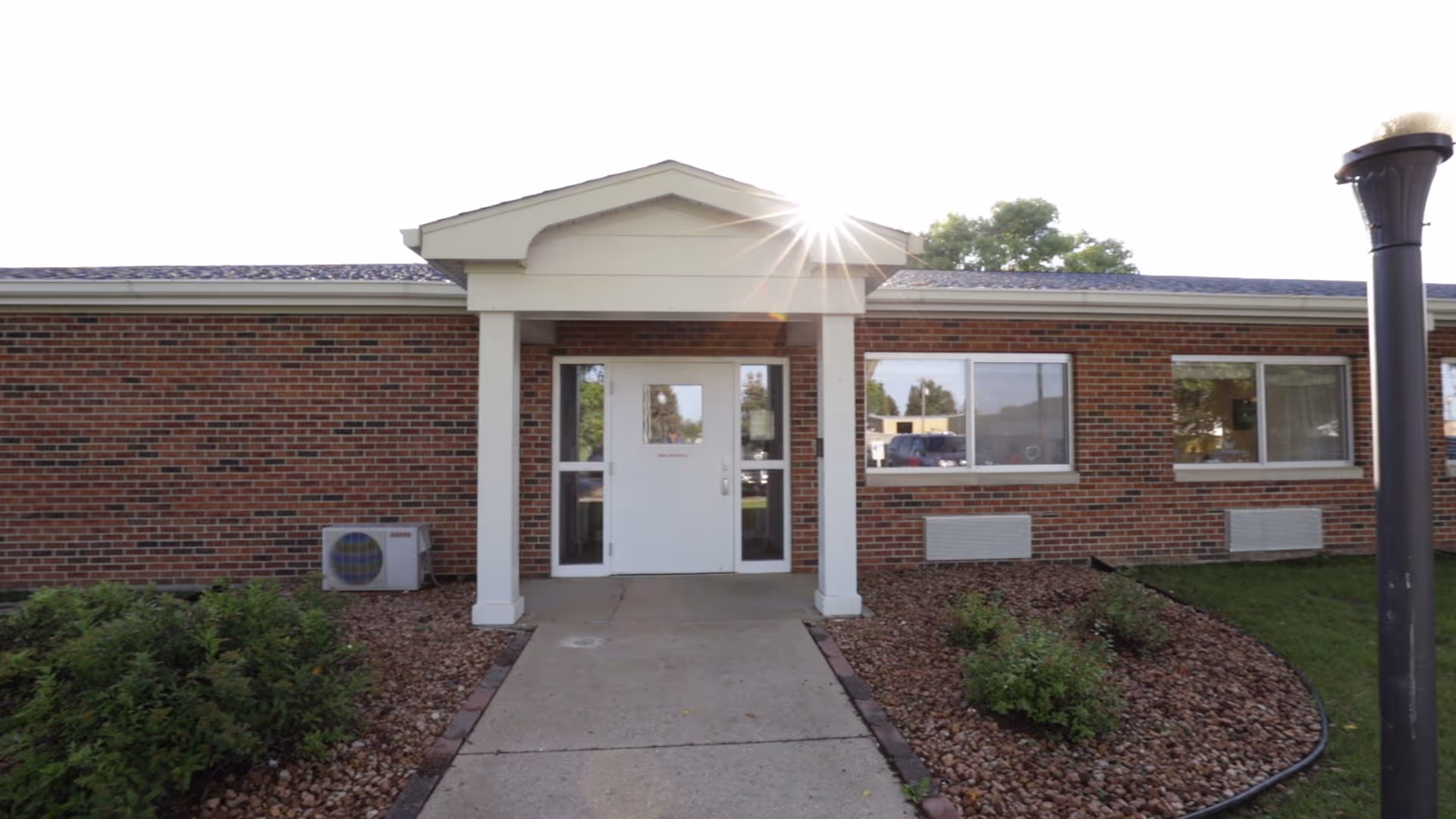Single-story brick building entrance with a portico, concrete walkway, and landscaped rock beds under a bright sun.