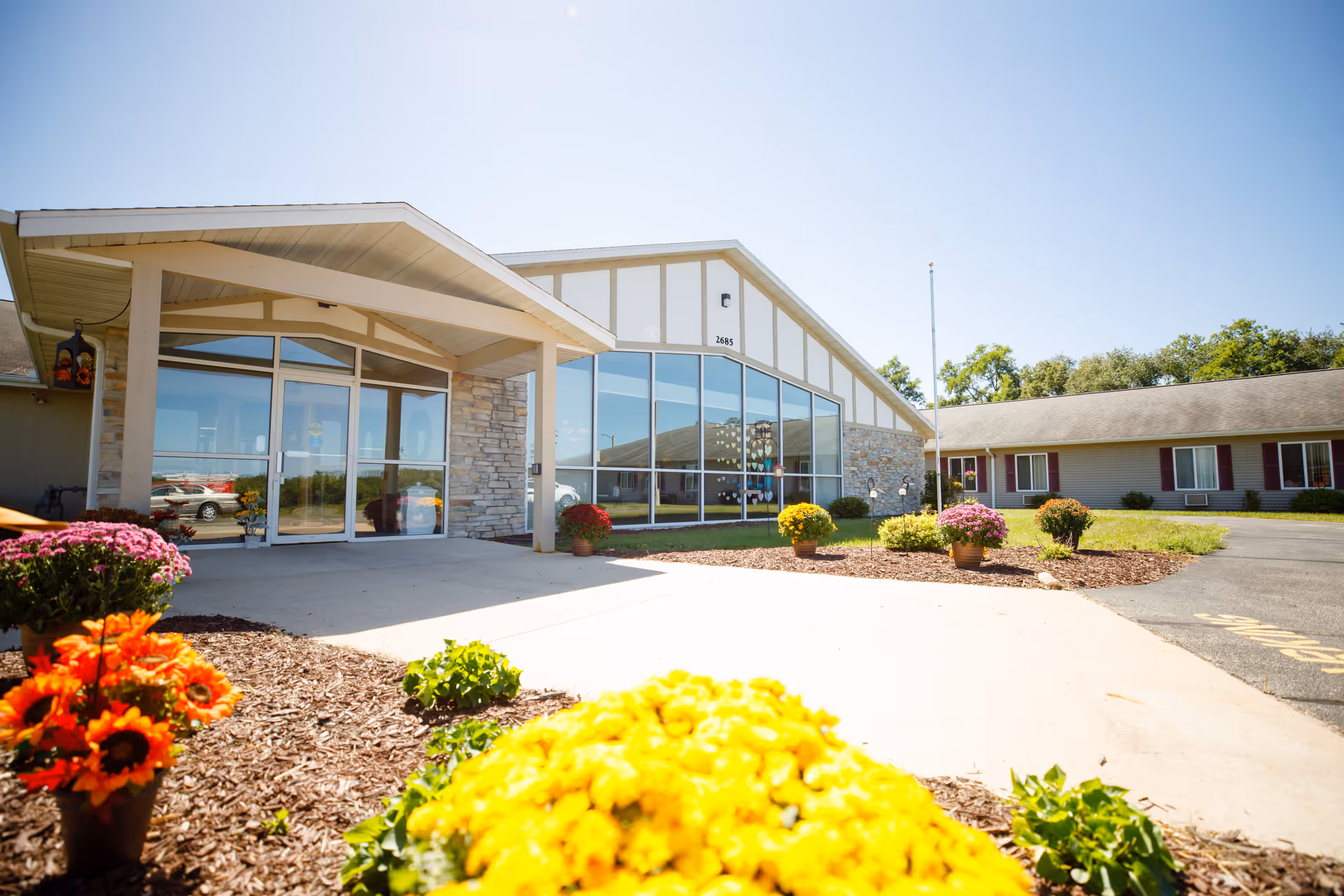 Exterior view of Heritage House of Portage Assisted Living facility on a sunny day, showing the entrance with large glass doors and windows, stone accents on the building, a paved walkway, and colorful flower pots with yellow, orange, and purple flowers in the landscaped garden area.