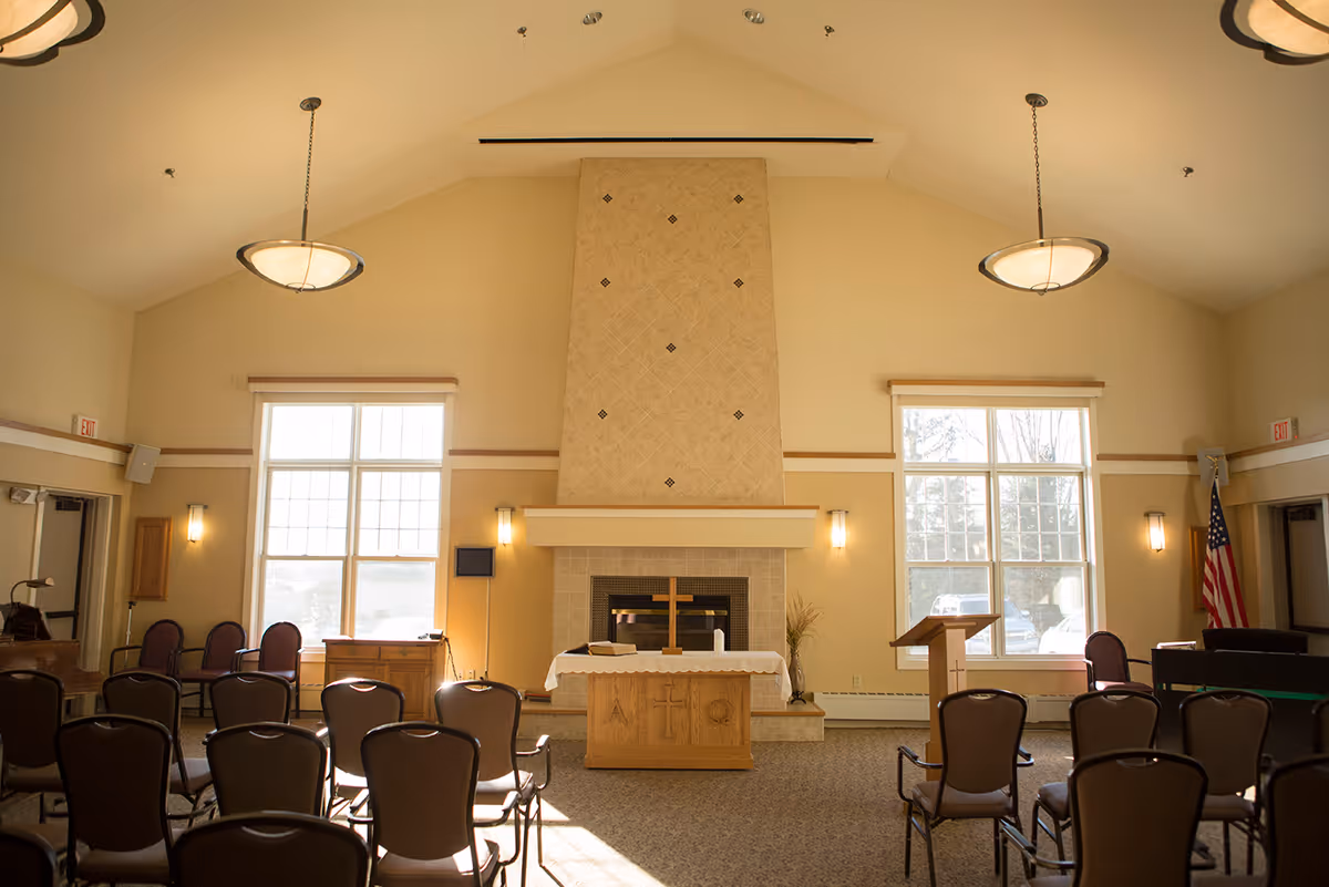 Interior view of a chapel or meeting room with rows of chairs facing a wooden altar with a cross, a fireplace behind it, large windows on either side, and an American flag in the corner.