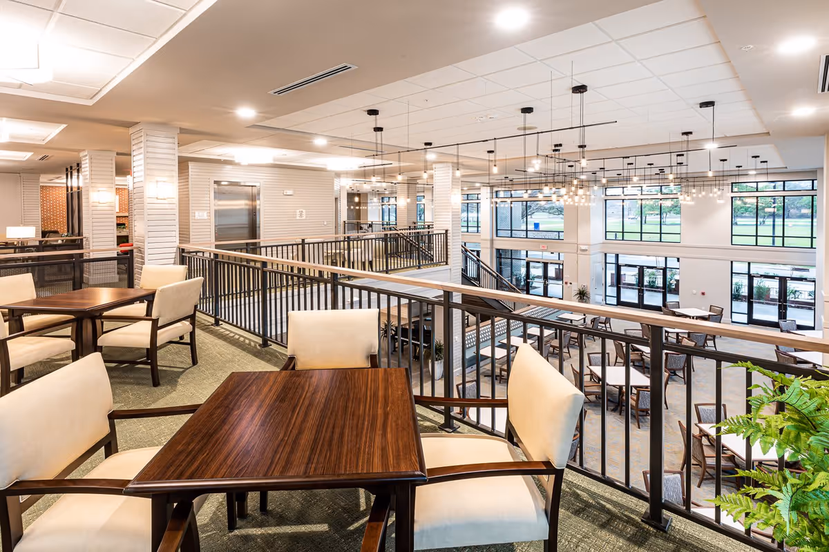 Interior view of a spacious senior living facility dining area with multiple tables and cushioned chairs. The area has large windows allowing natural light to fill the space, modern hanging light fixtures, and a railing overlooking a lower level with additional seating.