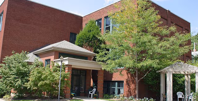 Front entrance of a red-brick retirement residence with a covered porch, trees, and outdoor chairs.