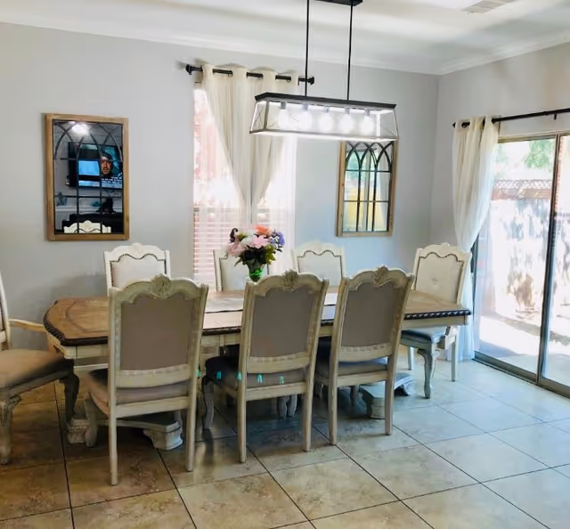 Bright dining room with a long wooden table surrounded by upholstered chairs, a rectangular chandelier, mirrors on the wall, and sliding glass doors with curtains.