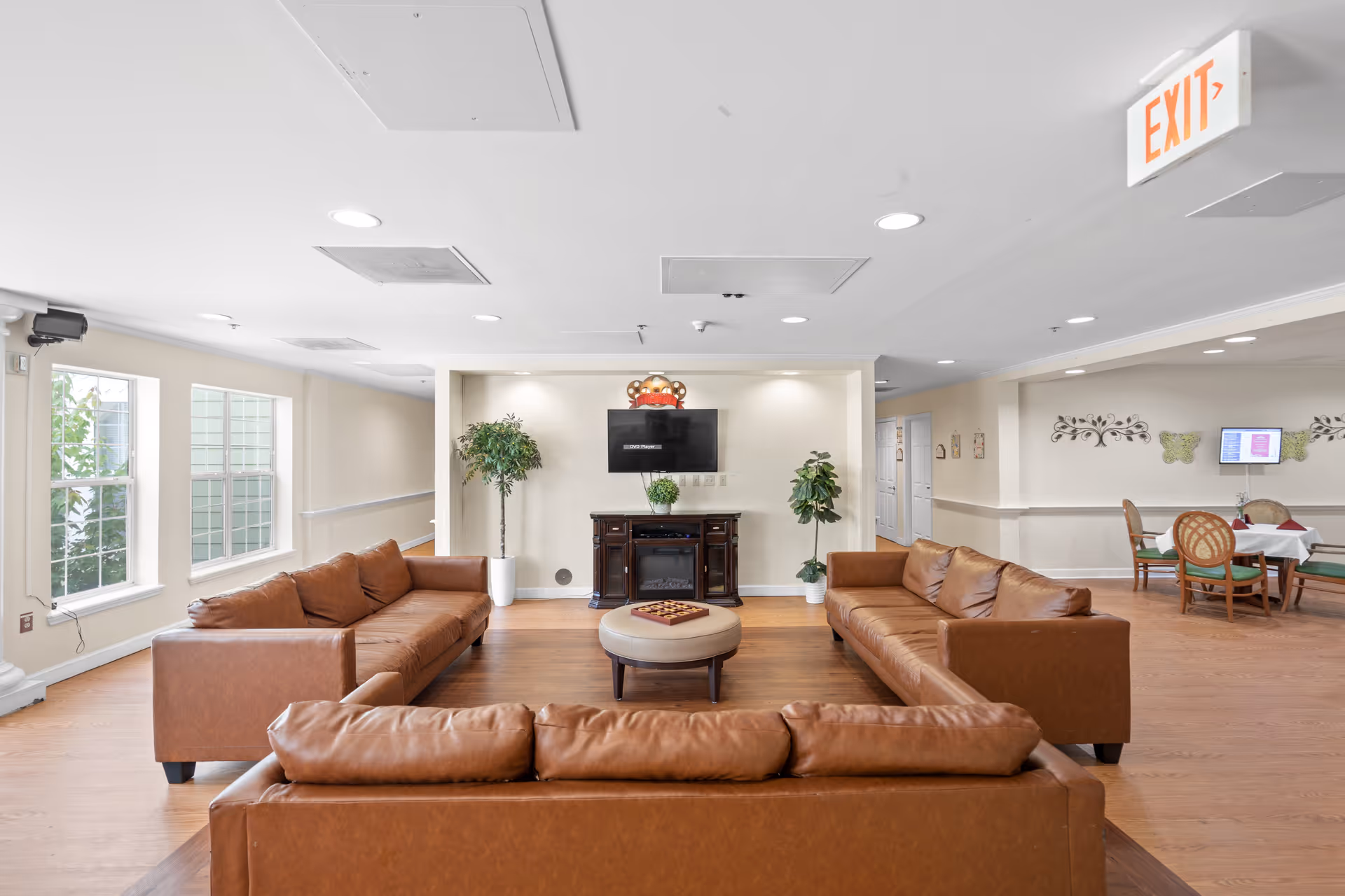 A bright and spacious common living area with three brown leather sofas arranged around a round ottoman. A flat-screen TV is mounted on the wall above a dark wooden cabinet, flanked by two potted plants. Large windows on the left side let in natural light. In the background, there is a dining area with tables and chairs, and an exit sign is visible on the ceiling.