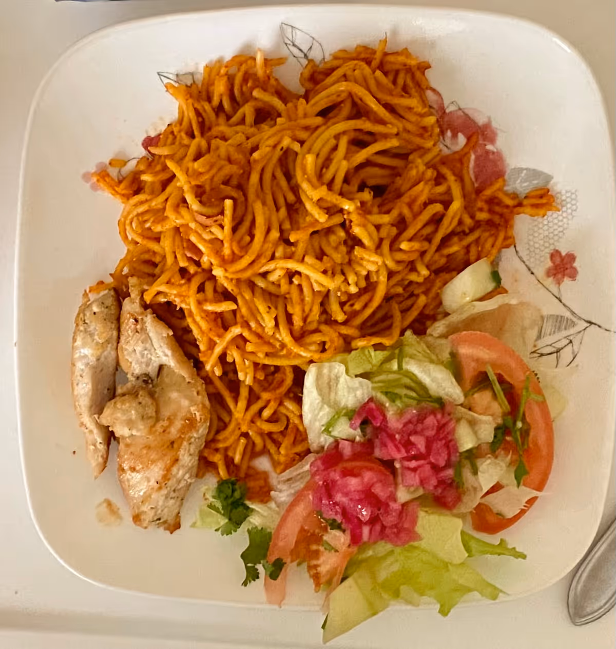 A white square plate with a serving of cooked spaghetti noodles in red sauce, two pieces of grilled chicken, and a side salad consisting of lettuce, tomato slices, and pickled red onions.