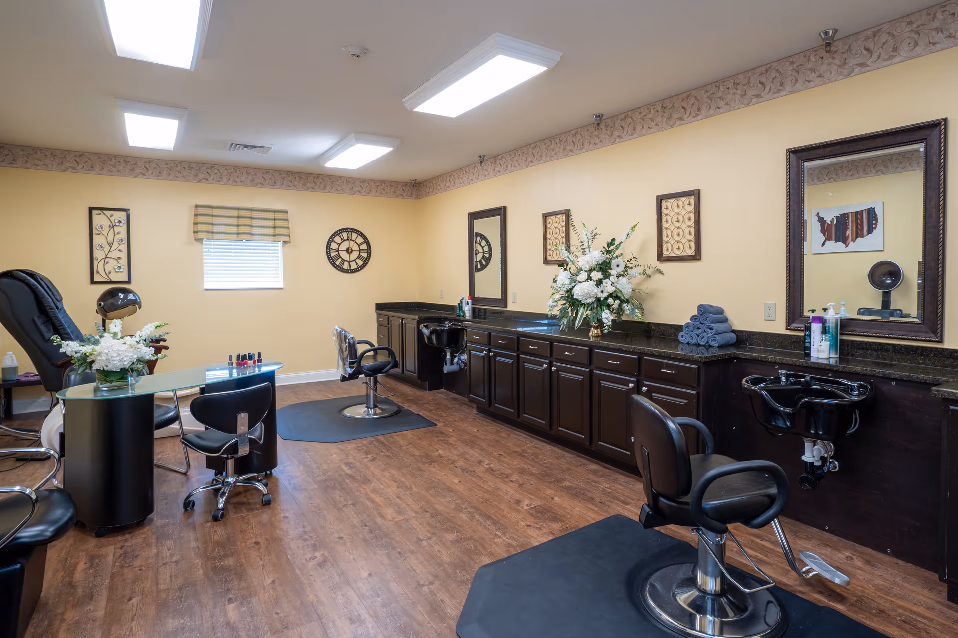 Interior view of a salon room with wooden flooring, two black salon chairs in front of sinks, a manicure table with chairs, decorative wall art, a clock, and floral arrangements on the countertop under large mirrors.