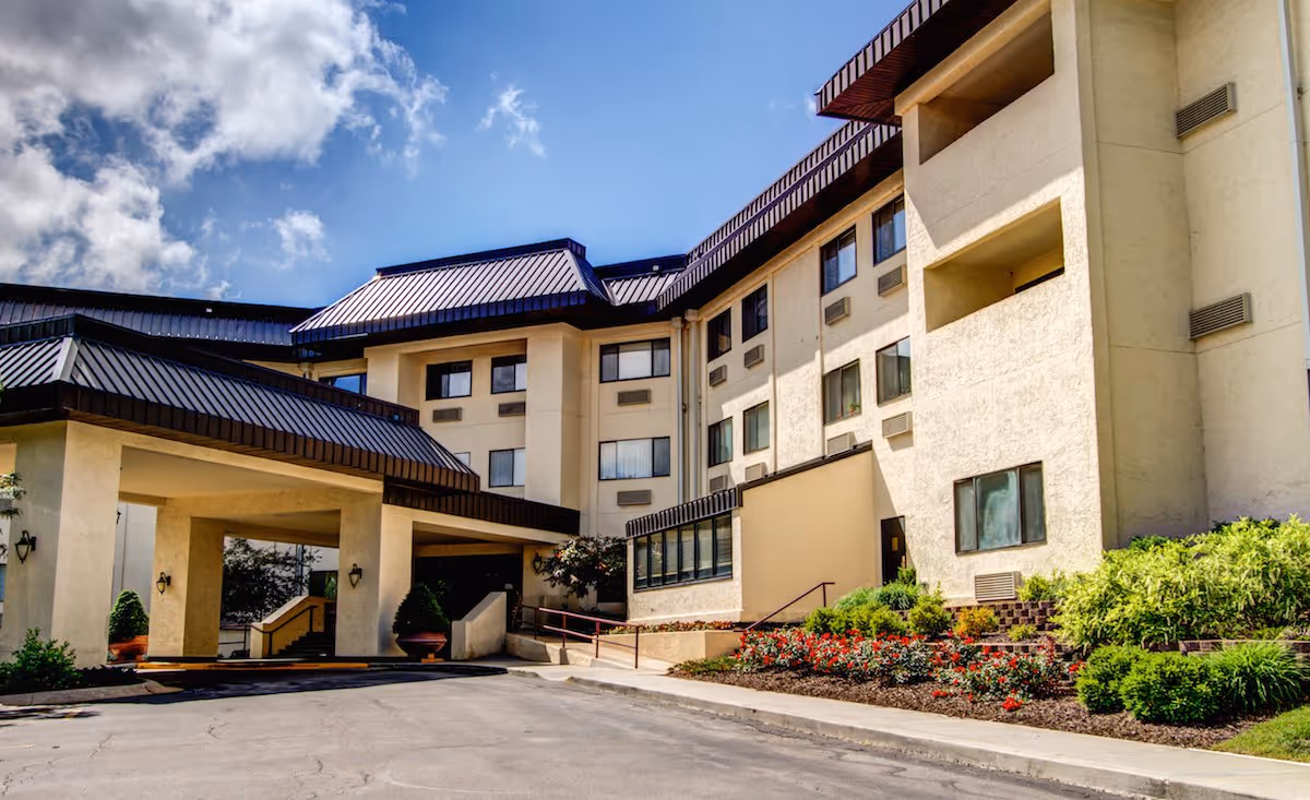 Exterior view of a multi-story senior living facility building with beige walls, dark brown roof accents, and a covered entrance. The foreground shows a paved driveway and landscaped flower beds with green shrubs and red flowers under a partly cloudy blue sky.