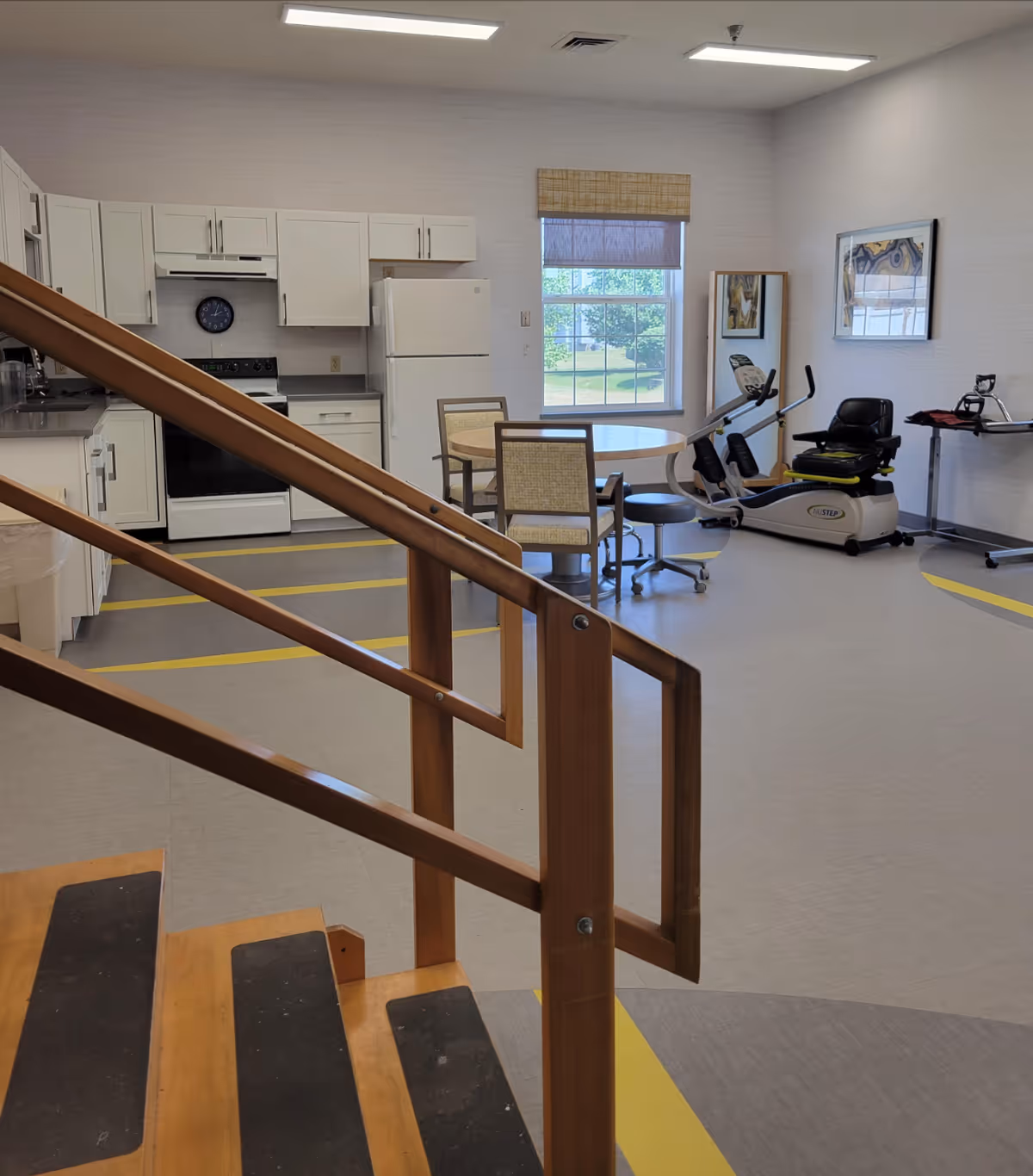 Interior view of a senior living facility room featuring a small kitchen area with white cabinets, a stove, and a refrigerator. In the foreground, there is a wooden handrail and steps. The room also includes a round table with chairs, an exercise machine, a stool, a full-length mirror, and framed artwork on the walls. A window with a bamboo shade lets in natural light.