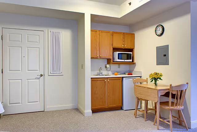 Small kitchenette area with wooden cabinets, a microwave, a mini refrigerator, and a sink. Next to the kitchenette is a small wooden dining table with two chairs and a vase of yellow flowers on top. To the left is a white door and a small window with a curtain. The walls are light-colored and the floor is carpeted.