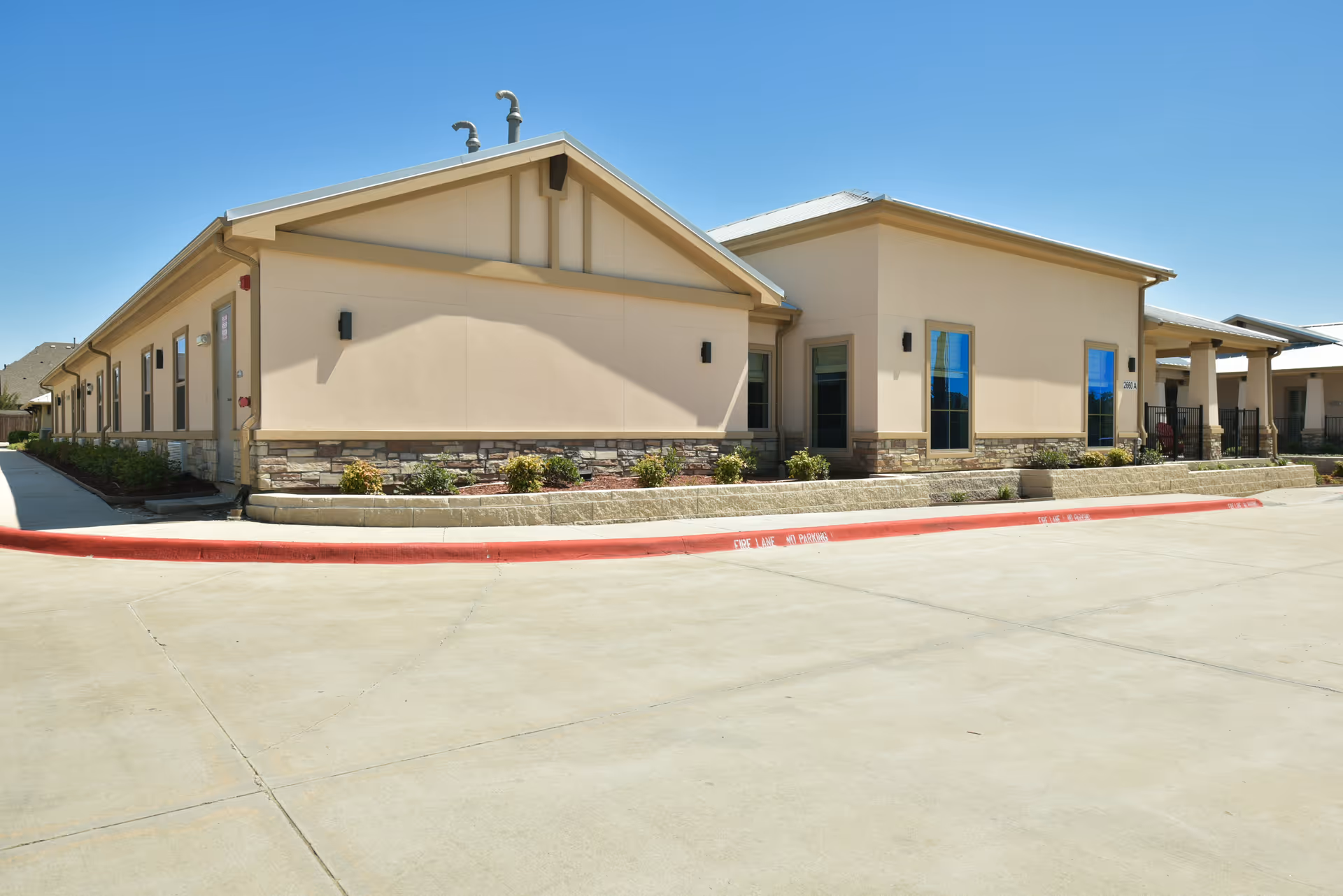 Single-story beige senior living facility building with stone trim, windows, and a red fire-lane curb under a clear blue sky.