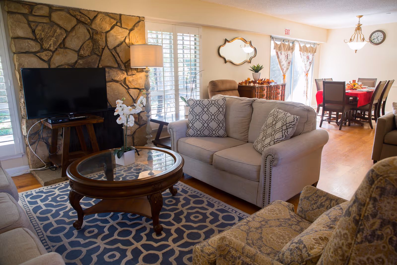 A cozy living room with a beige loveseat adorned with patterned pillows, a glass-top wooden coffee table with a white orchid plant, a stone fireplace with a flat-screen TV on a wooden stand, a floor lamp, and a patterned armchair. In the background, there is a dining area with a wooden table covered with a red tablecloth and six chairs, floral curtains on sliding glass doors, a wall clock, and a decorative mirror above a wooden sideboard.