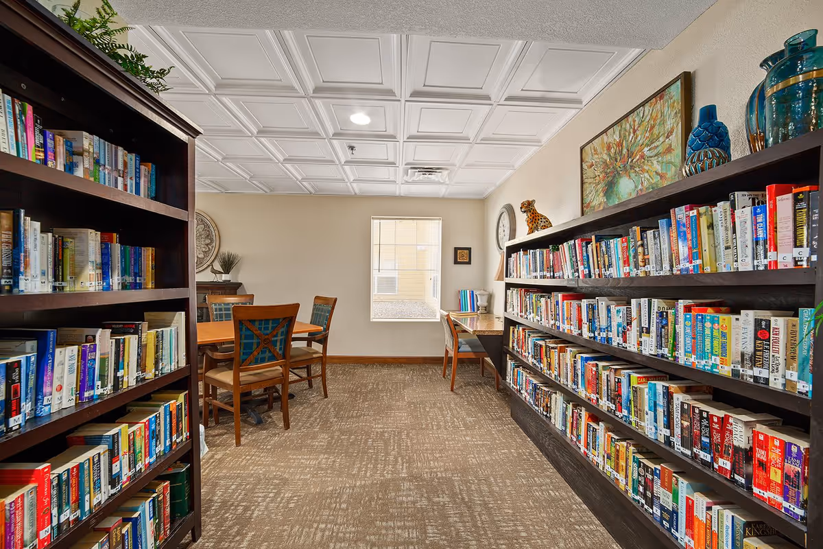 Interior of a library room in a senior living facility with two large dark wooden bookshelves filled with books on either side. There is a wooden table with four chairs in the center and a desk with a chair near a window at the back. The room has a beige carpet, a white coffered ceiling, and decorative items including a clock, a painting, and vases.