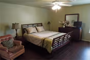 A cozy bedroom with a wooden bed frame, beige bedding, a wooden dresser with a TV on top, a side table with a lamp, a comfortable armchair with a cushion, and a ceiling fan with lights. The room has wooden flooring and light-colored walls.