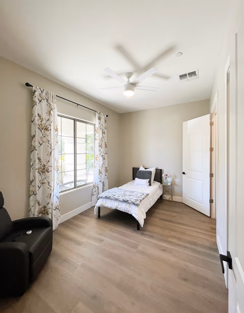 A bright bedroom with light-colored walls and wood flooring. The room features a single bed with white bedding and gray pillows, a small side table with a decorative item, a large window with floral curtains, a ceiling fan with a light, and a black recliner chair.