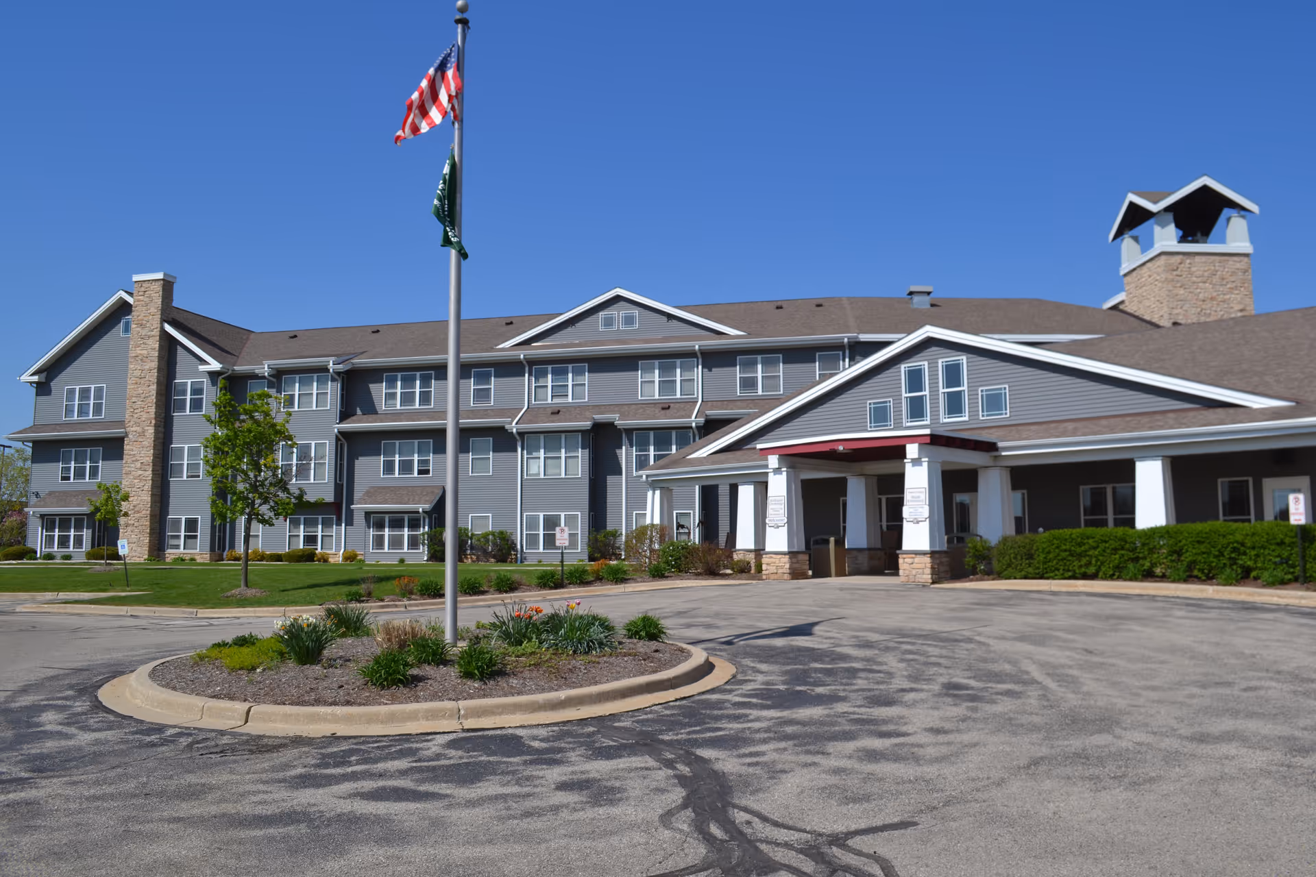 Exterior view of a large senior living facility building with multiple windows, a covered entrance, and a circular driveway with a landscaped island featuring plants and two flagpoles flying the American flag and another flag, under a clear blue sky.