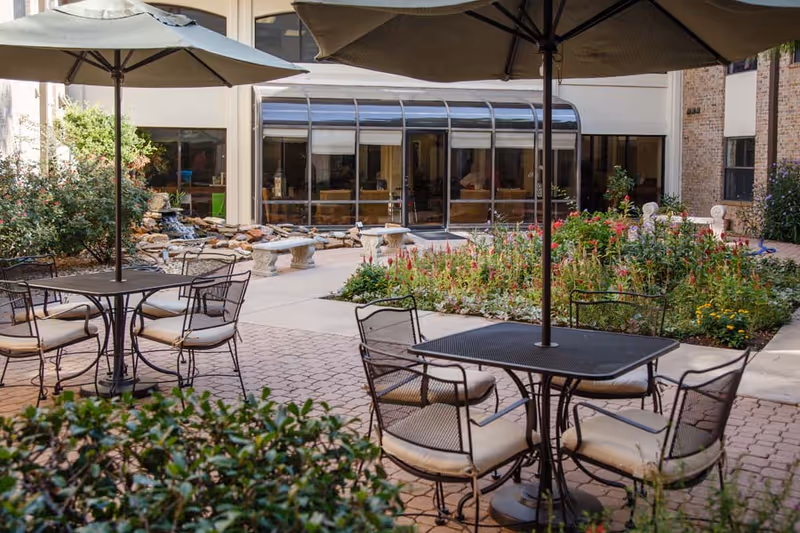 Outdoor courtyard with patio tables and umbrellas, planted flower beds and a small fountain in front of a building's glass-enclosed common area.
