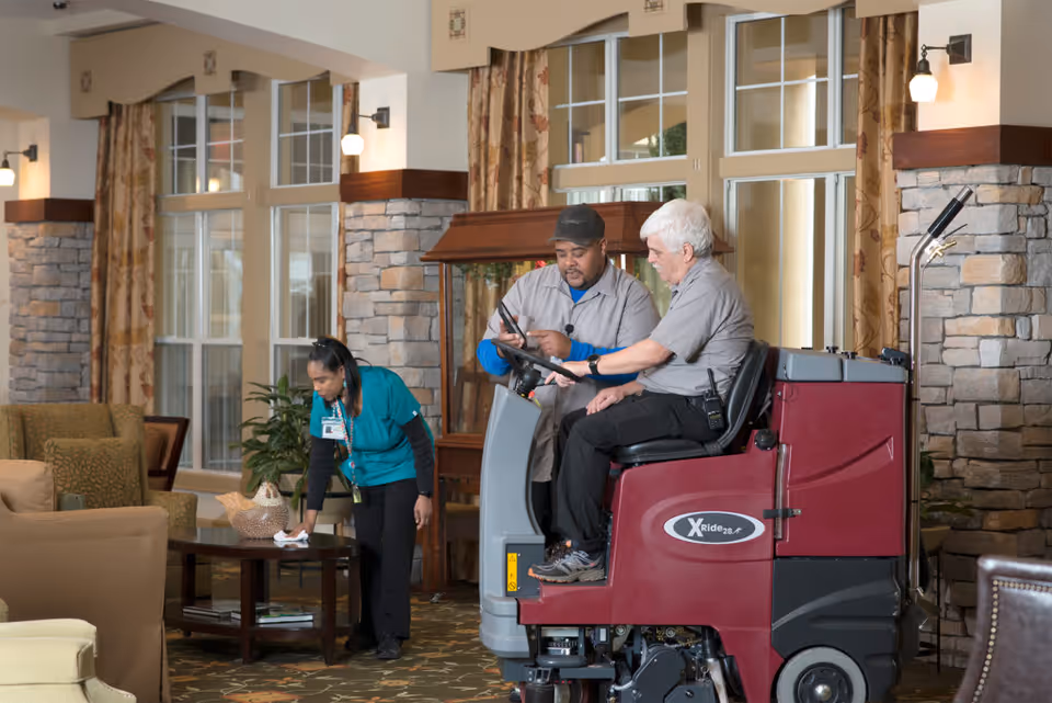 Two maintenance workers operating a floor cleaning machine inside a well-lit common area with stone pillars and large windows. A staff member in teal scrubs is cleaning a wooden coffee table nearby. The room has comfortable seating and patterned curtains.