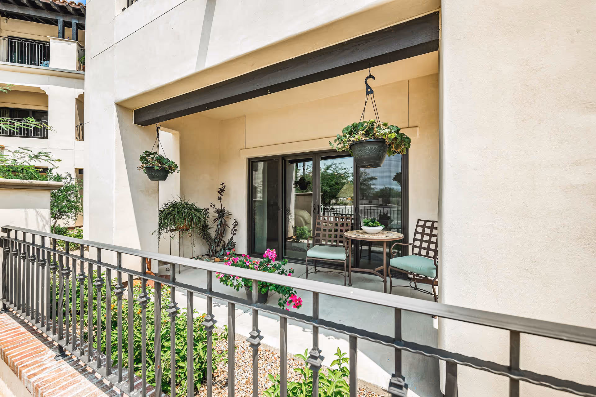 Small covered patio with hanging plants, a round table and two chairs in front of sliding glass doors behind a decorative metal railing.