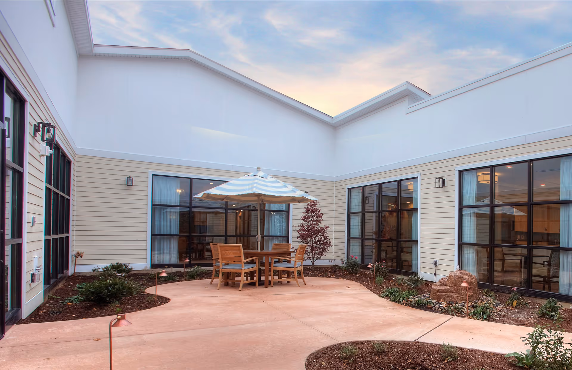 Enclosed courtyard with a patio table, umbrella and chairs surrounded by windows and landscaping.