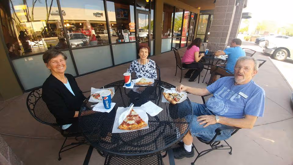 Three older adults sitting around a round outdoor metal table eating pizza and drinking soda outside a storefront.