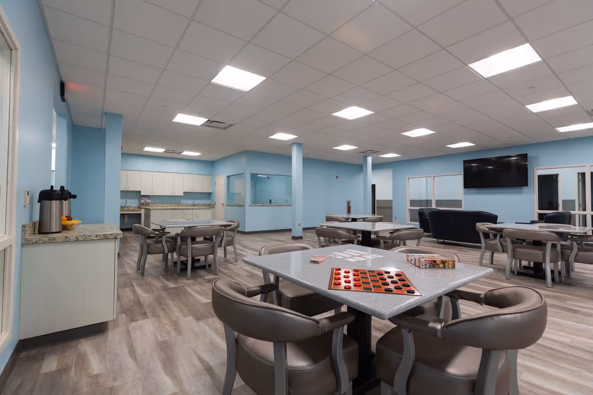 A spacious common area with light blue walls and wood-patterned flooring. Several tables with chairs are arranged throughout the room, some with board games like checkers and cards on them. In the background, there is a kitchenette with white cabinets and a countertop. A large flat-screen TV is mounted on the wall above a dark blue couch and chairs. The ceiling has recessed lighting panels.